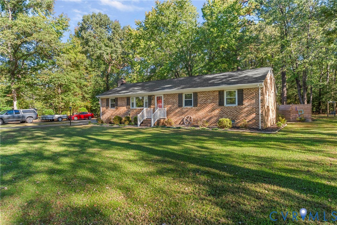 Single-story brick exterior featuring a shingled gable roof, expansive lawn, white railing entrance, and dense perimeter tree canopy