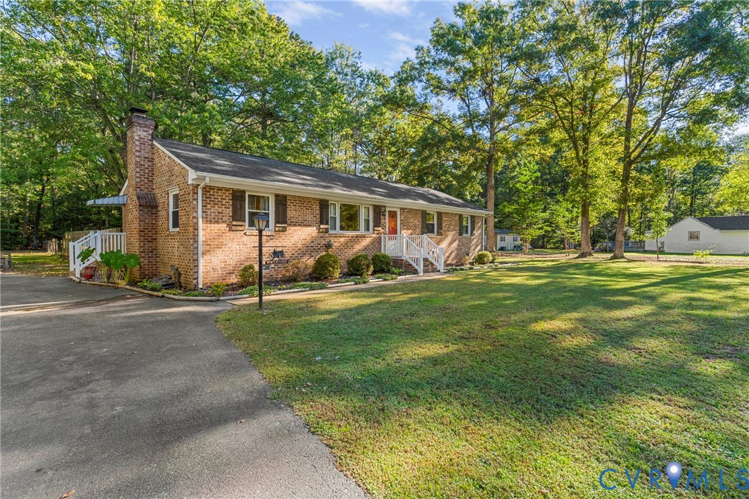 Brick exterior featuring a masonry chimney, dark-toned shutters, a paved driveway, and white railing accents, all situated on a lot with mature trees
