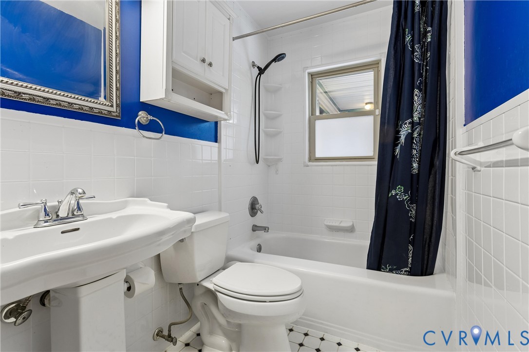 Pedestal sink with polished chrome faucet, white wall-mounted storage cabinet, white subway-style wall tile, built-in tub and shower combination, and geometric tile flooring