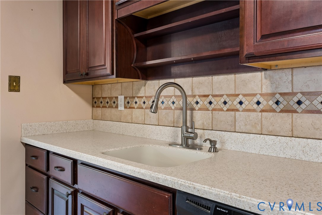 Dark wood-finish cabinetry featuring open shelving, light-toned composite countertop, integrated sink with a stainless steel gooseneck faucet, and decorative tile backsplash with diamond-pattern accents