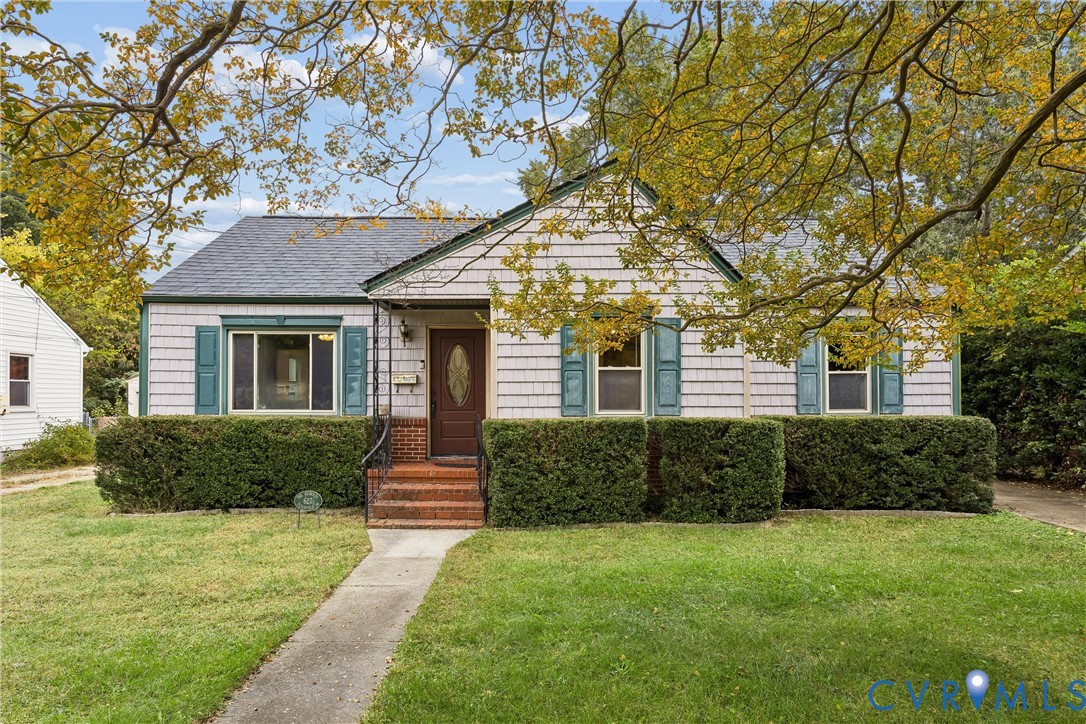 Single-story residence featuring light-colored shingle siding, decorative green shutters, a central entrance with a brick staircase, and a concrete walkway Single-story residence featuring light-colored shingle siding, decorative green shutters, a central entrance with a brick staircase, and a concrete walkway