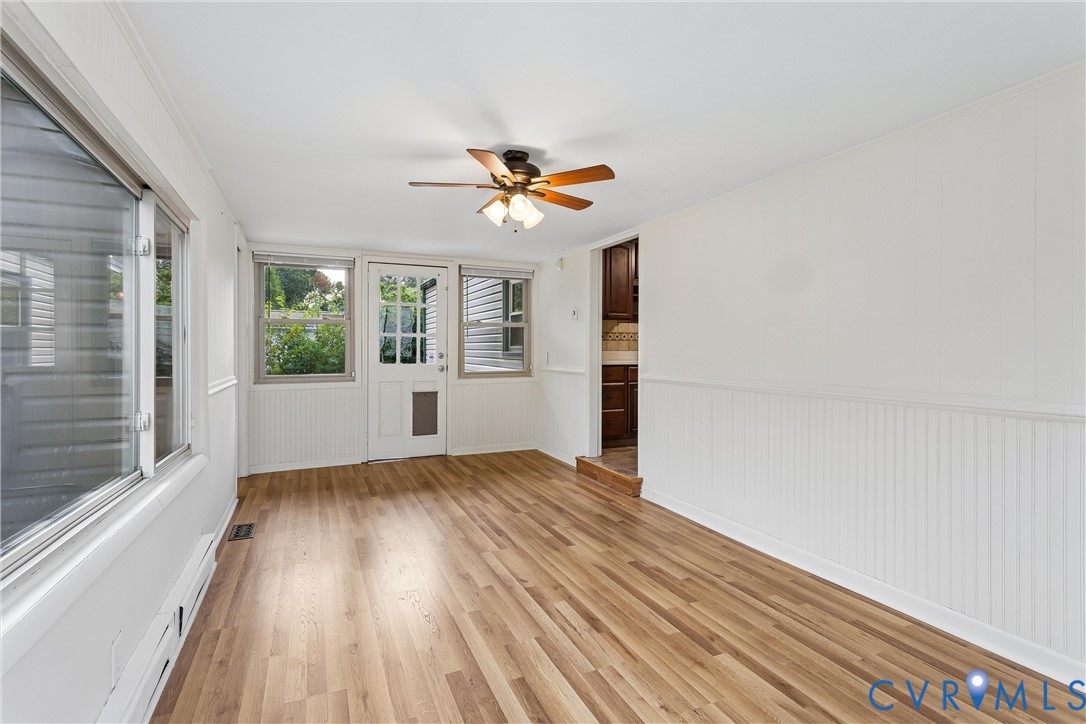 Wood-finish flooring, white beadboard wainscoting, multiple windows, and a multi-light exterior door with a ceiling fan