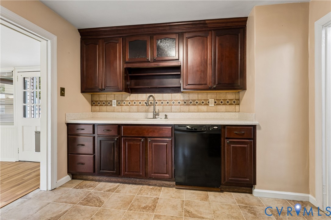 Dark wood cabinetry with glass-front accents, solid surface countertops, patterned ceramic tile backsplash, integrated undermount sink with gooseneck faucet, and stone-finish tile flooring