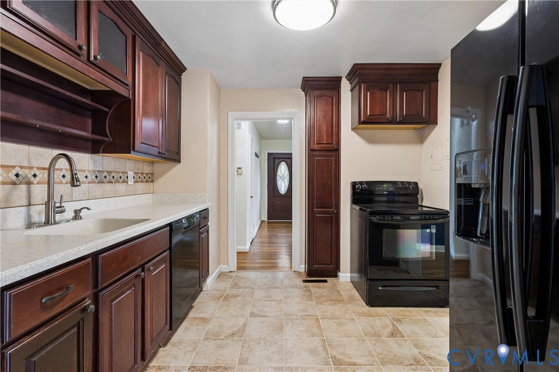 Kitchen featuring dark-toned cabinetry, solid surface countertops, tile backsplash with decorative inlay, built-in floor-to-ceiling pantry storage, and black appliance suite