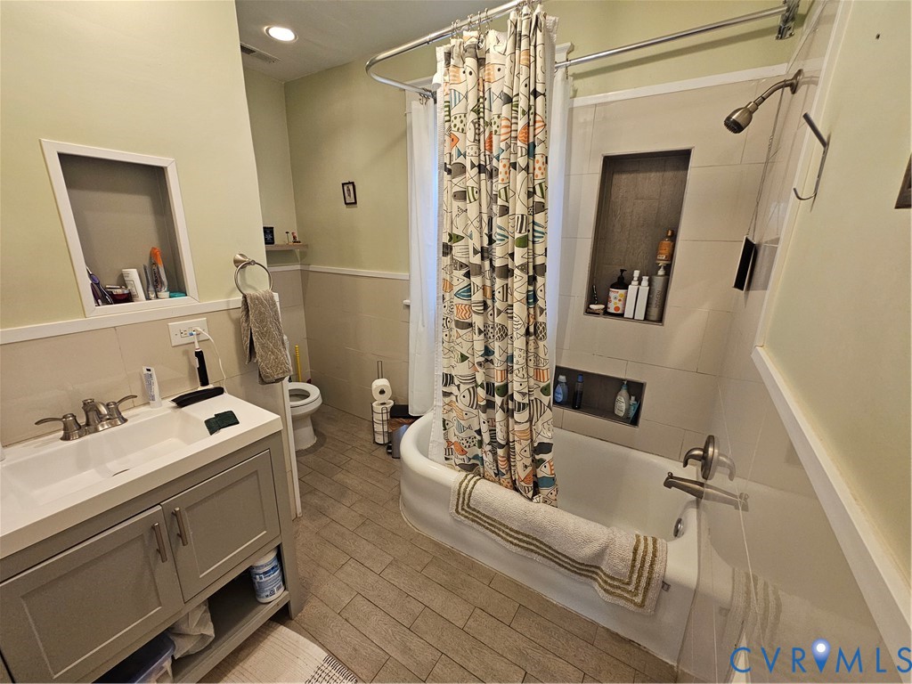 Bathroom featuring a gray vanity with integrated sink, tile-surround tub with dual recessed niches, wood-finish flooring, and wainscoting