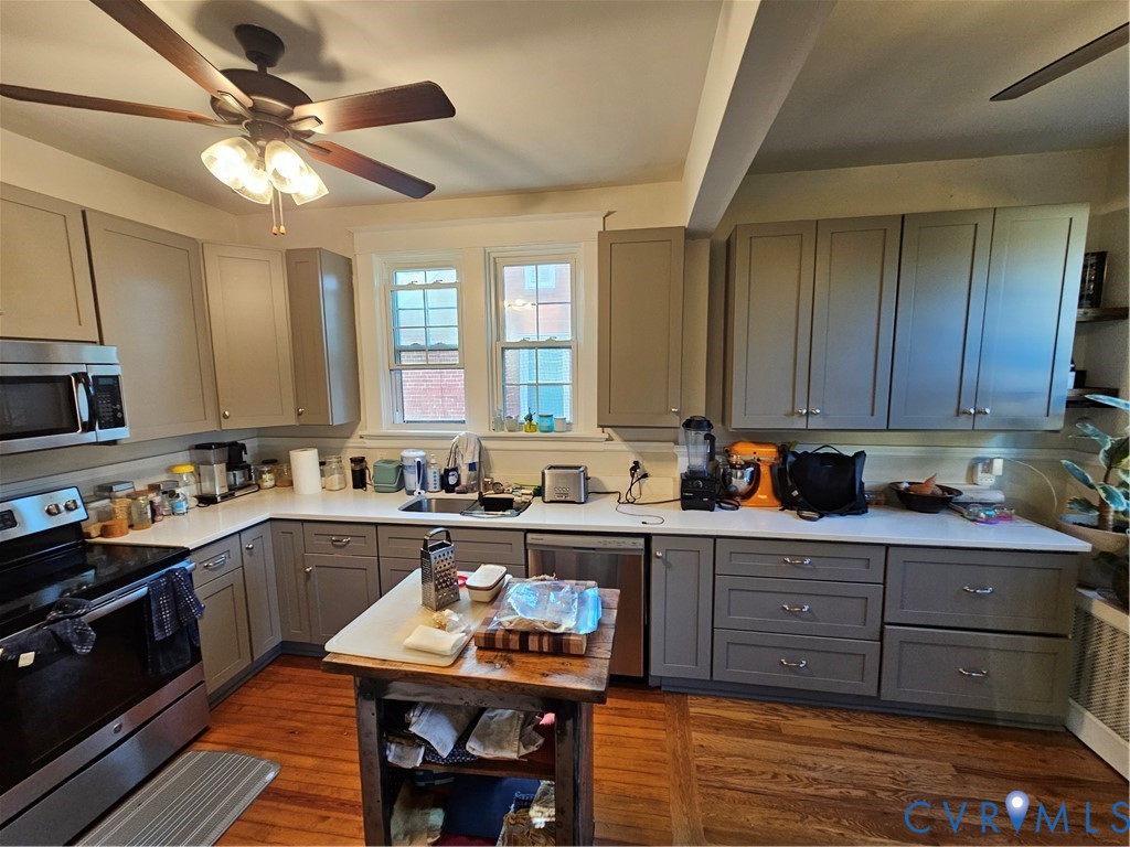 Kitchen featuring shaker-style cabinetry, solid-surface countertops, stainless steel appliances, wood flooring, and a ceiling fan with integrated lighting