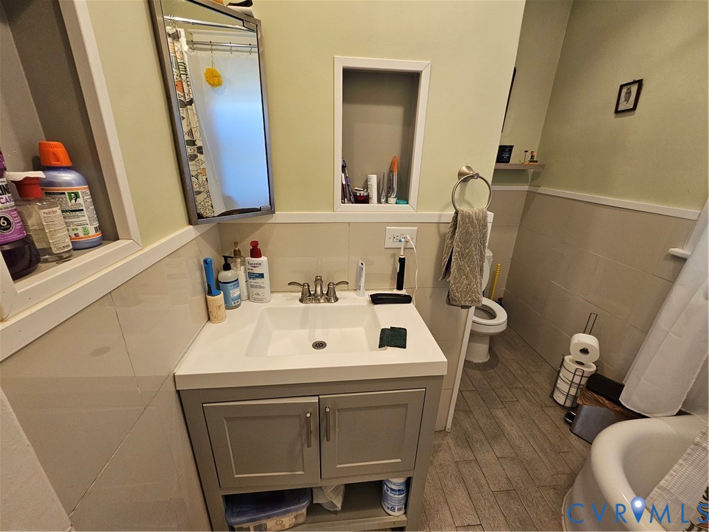 Bathroom featuring a vanity with a white integrated sink, polished metal faucet, inset wall shelving, partial wall tiling, and wood-finish flooring