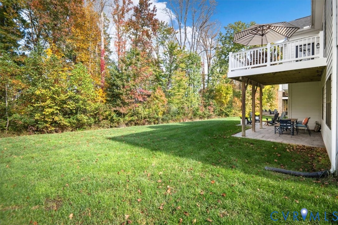 Rear exterior featuring an elevated wood deck with white railings, a paved stone patio, and an expansive lawn bordered by mature trees