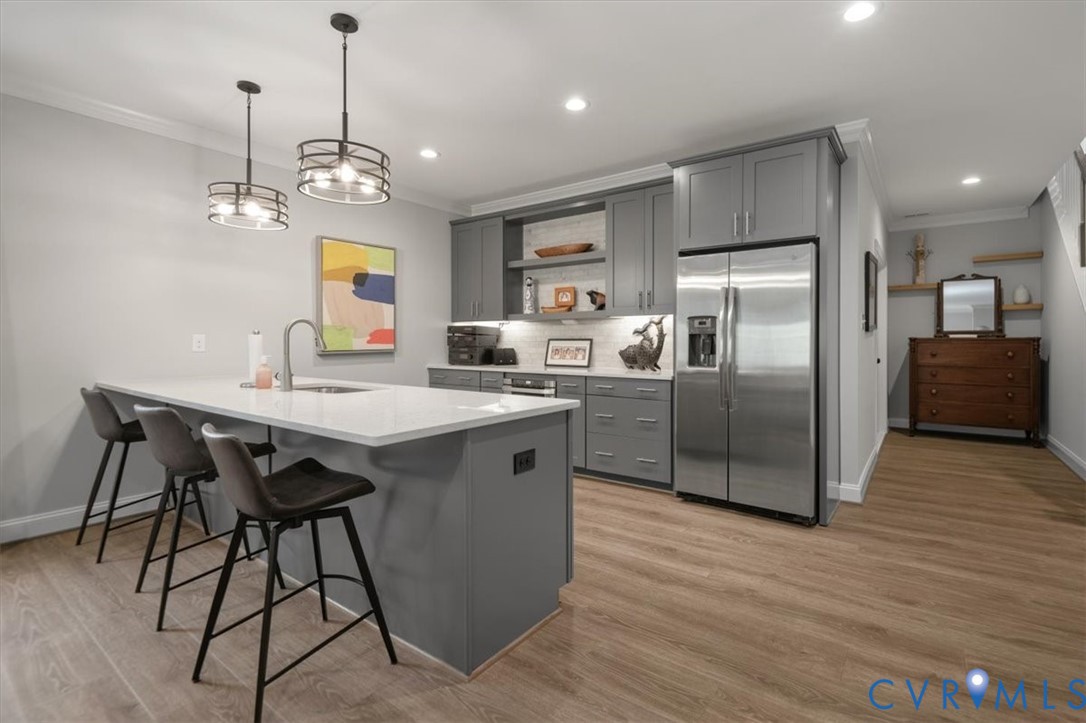 Terrace Level Kitchen featuring a central island with a stone countertop and integrated sink, gray cabinetry, stainless steel refrigerator, wood-finish flooring, and dual hanging pendant light fixtures