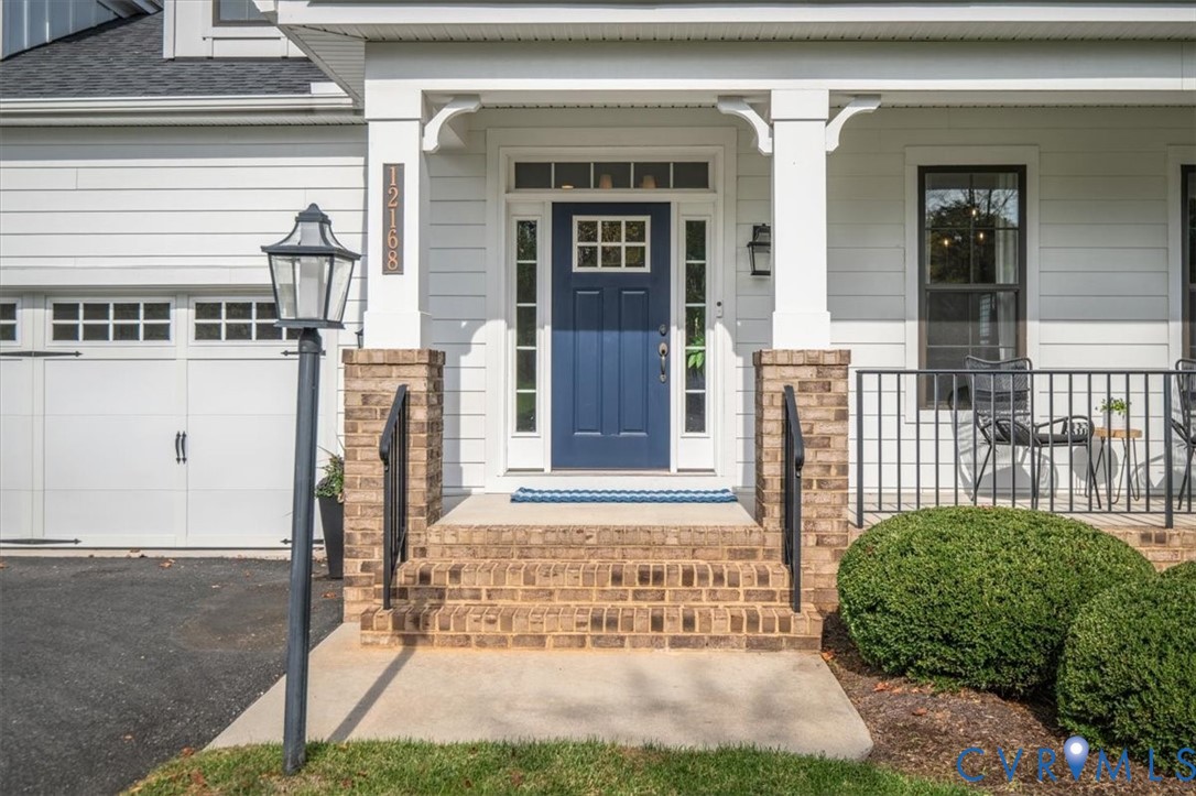 Exterior entry featuring horizontal siding, a covered porch with brick foundation piers, a solid-color paneled door with sidelights and transom window, and a carriage-style garage door