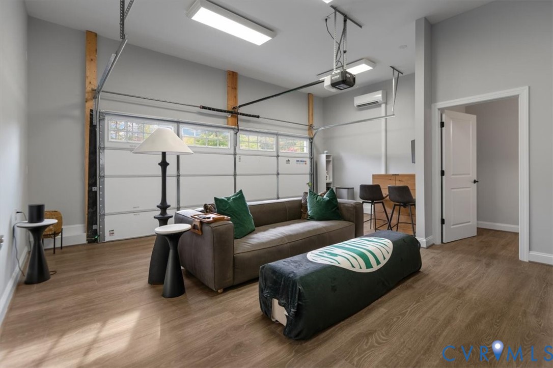 Finished Garage space featuring wood-finish flooring, exposed timber structural posts, white sectional overhead door with glass inserts, wall-mounted ductless climate control unit, and overhead rectangular LED lighting