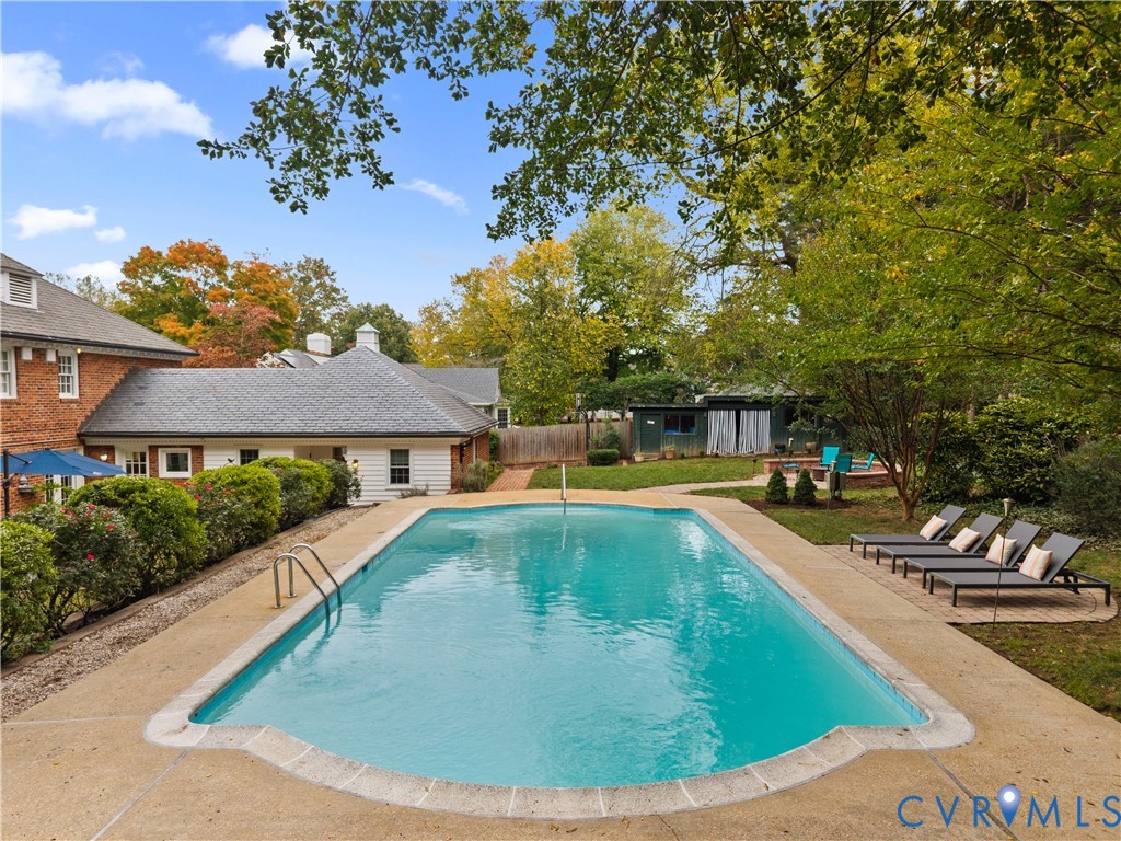 In-ground swimming pool with stone coping, expansive concrete pool deck, mature trees, and brick exterior residence