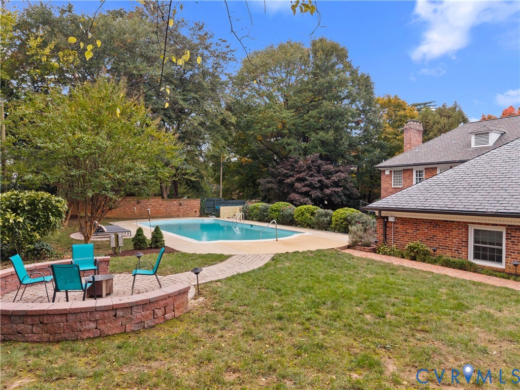 Backyard featuring a swimming pool with concrete decking, a circular paver fire pit area, a brick retaining wall, and mature landscaping