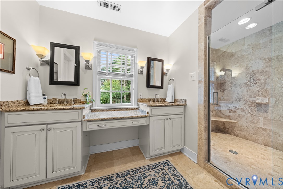 Double vanity with stone countertop, central built-in vanity desk, wall-mounted sconce lighting, and walk-in shower with stone tile surround and integrated bench