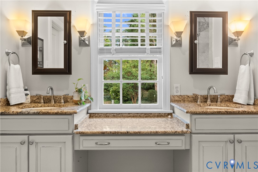 Double vanity featuring stone countertops, integrated cabinetry, dual wall-mounted mirrors, sconce lighting, and a center sitting area