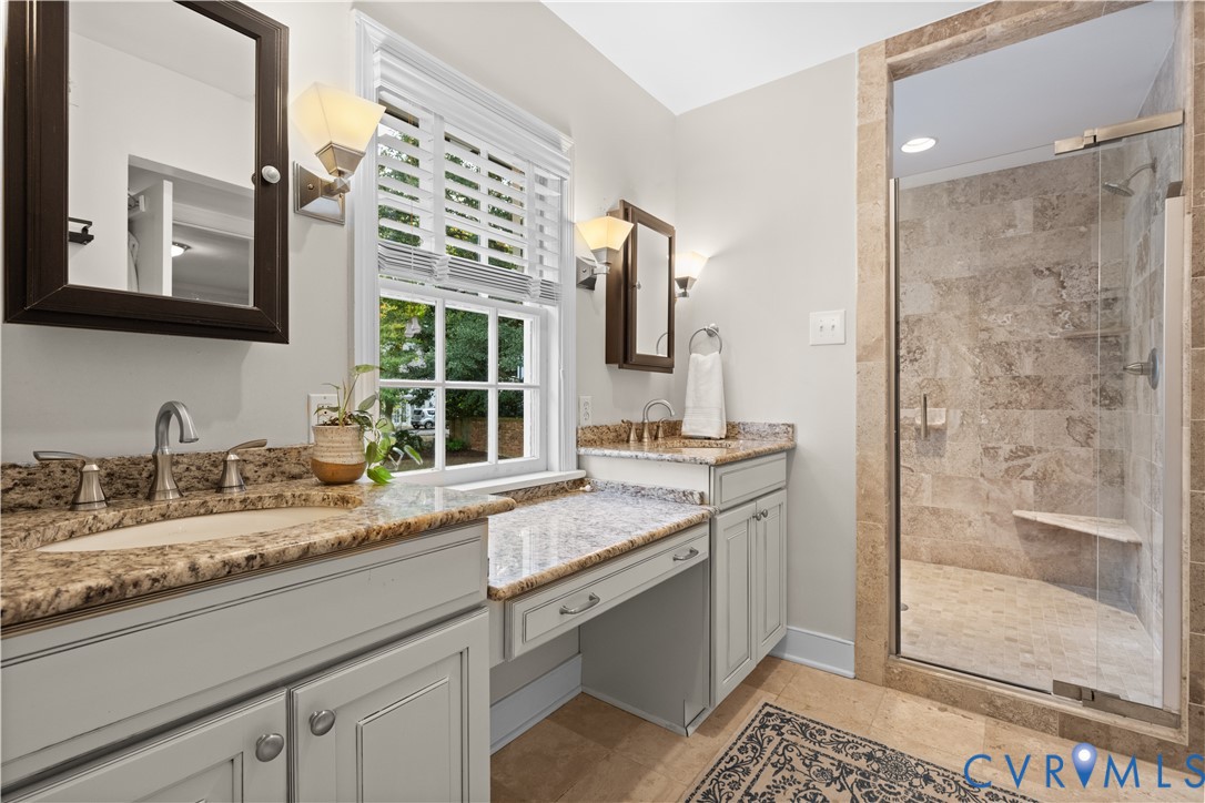Dual vanity with stone-surface countertops, walk-in shower featuring glass enclosure and stone-tile surround, and built-in vanity station with cabinetry