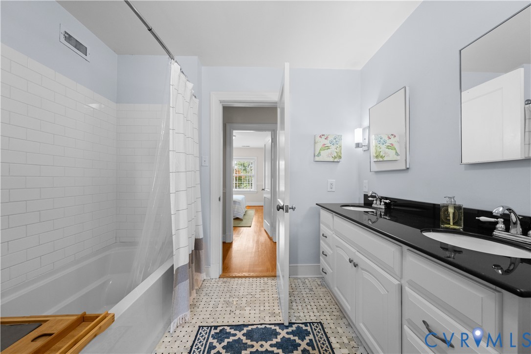 Bathroom featuring a dual-sink vanity with a dark stone countertop, white cabinetry, subway tile bathtub surround, and geometric patterned floor tiling