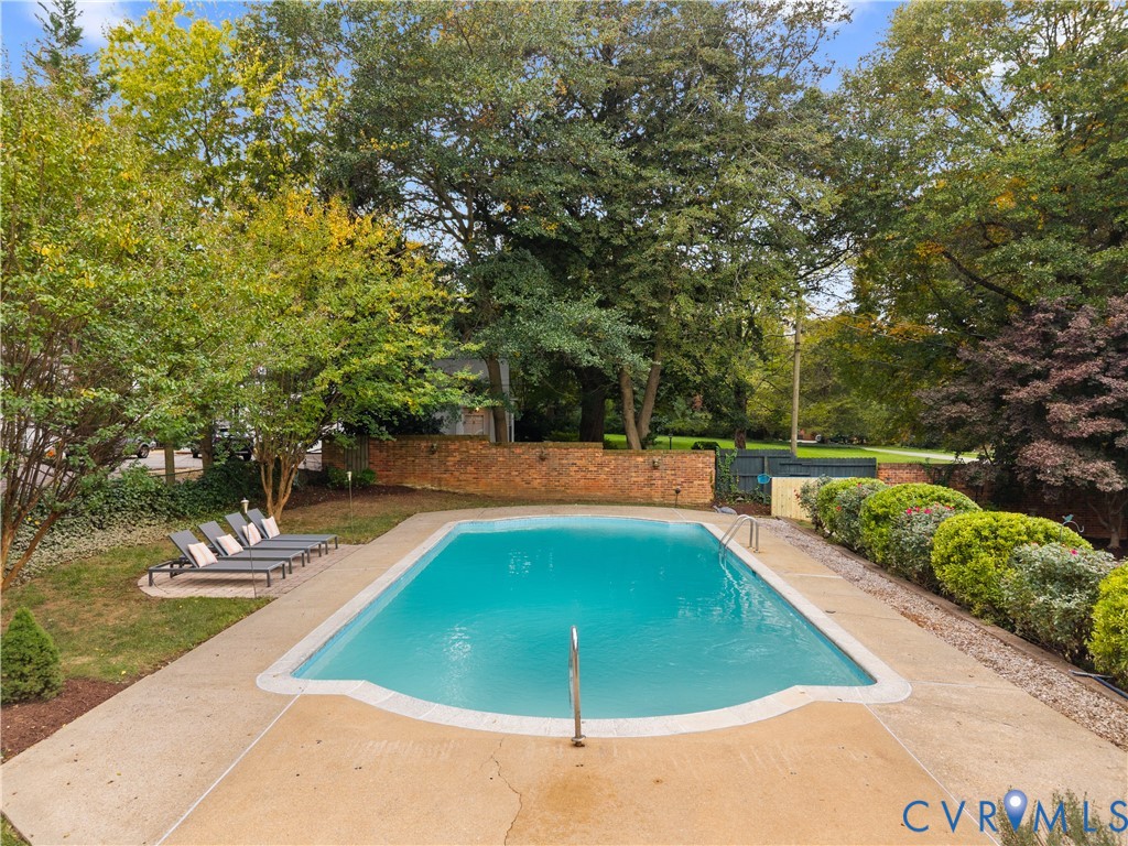 Inground swimming pool with metal ladder, concrete surround, brick privacy wall, and mature landscaping