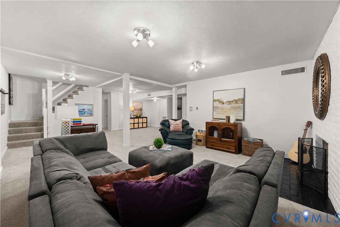 Open-concept interior featuring a painted brick fireplace, neutral carpeting, structural columns, and a carpeted staircase with white balusters