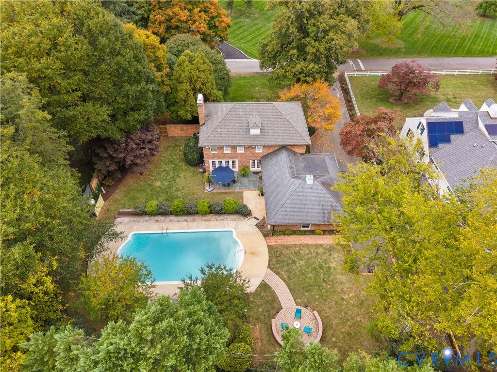Aerial view featuring a brick exterior residence, in-ground swimming pool, stone patio, brick-paved walkway, and a circular fire pit area set within a wooded lot