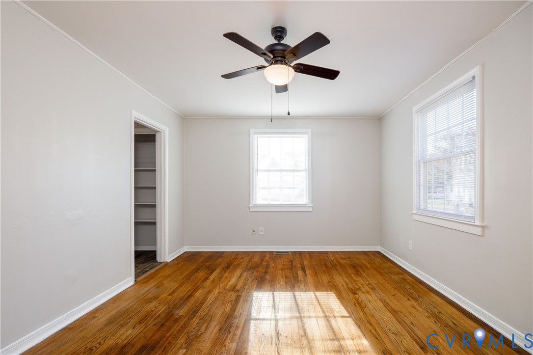 Room featuring wood-finish flooring, a ceiling fan with integrated lighting, two windows with white trim, and built-in shelving within a doorway