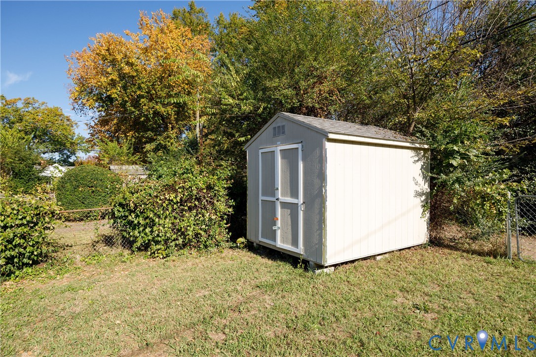 Outdoor storage structure featuring light-colored siding, double doors, and a gabled roof set on a level lawn with perimeter fencing