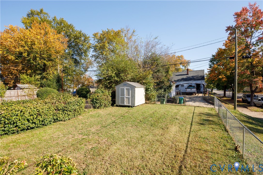 Backyard featuring a grass lawn, chain-link fencing, a detached storage shed, mature trees, and a gravel driveway