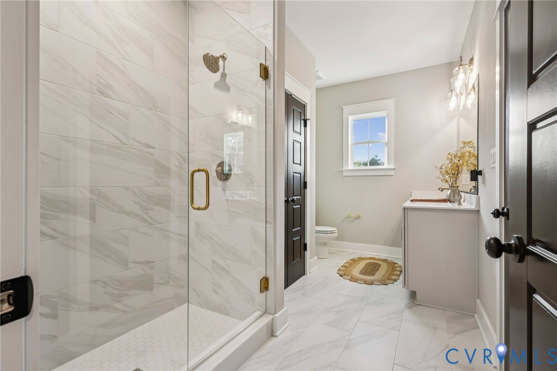 Spacious bathroom featuring a glass-enclosed shower with gold-tone fixtures, white tile flooring, a single vanity with a white countertop, and a window providing natural light