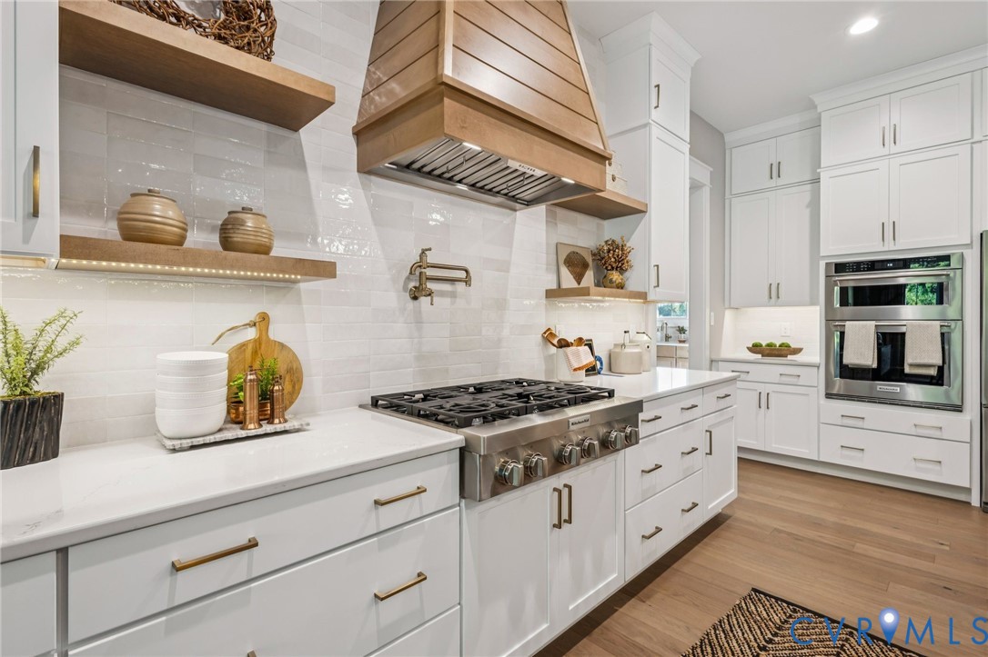 Contemporary kitchen featuring a wood-finish range hood, stainless steel gas range, double wall ovens, and white shaker cabinetry with brass hardware