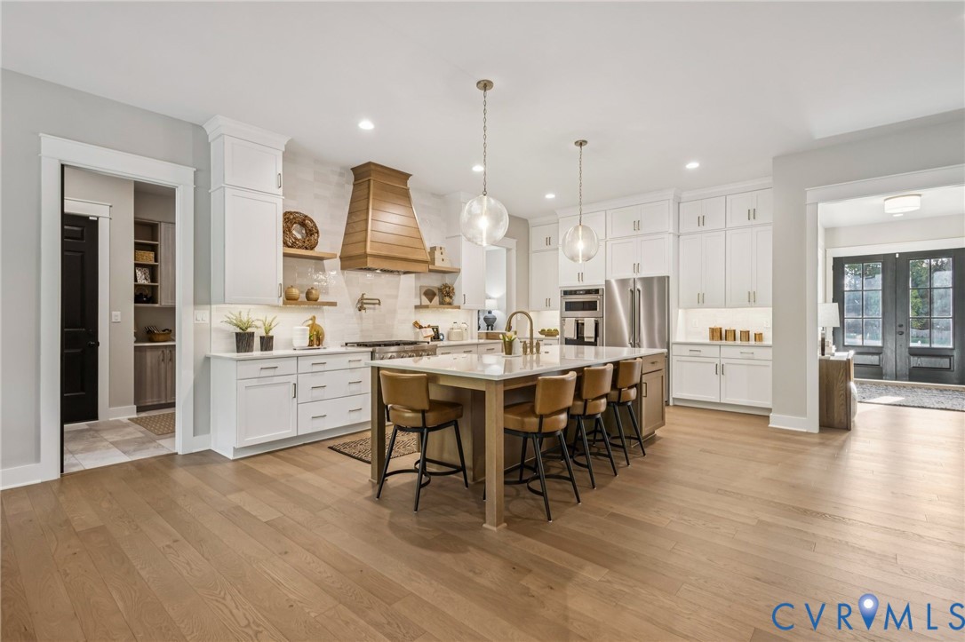 Expansive kitchen featuring a large central island, white cabinetry, integrated stainless steel appliances, a wood-finish range hood, and light wood-finish flooring