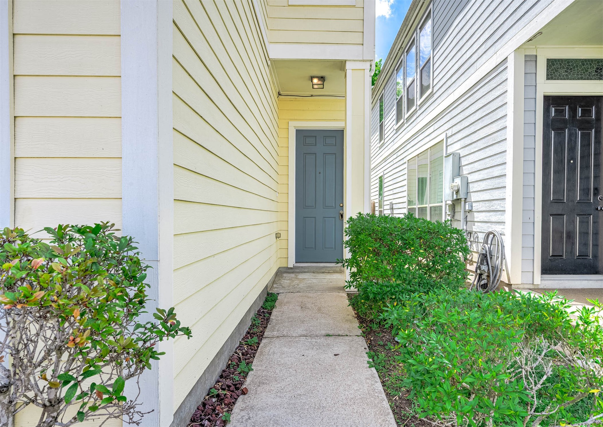 Welcome to your new home, featuring a charming blue door framed by pastel painted siding. Inviting Entrance with Lush green shrubs and grass. Welcome to your new home, featuring a charming blue door framed by pastel painted siding. Inviting Entrance with Lush green shrubs and grass.