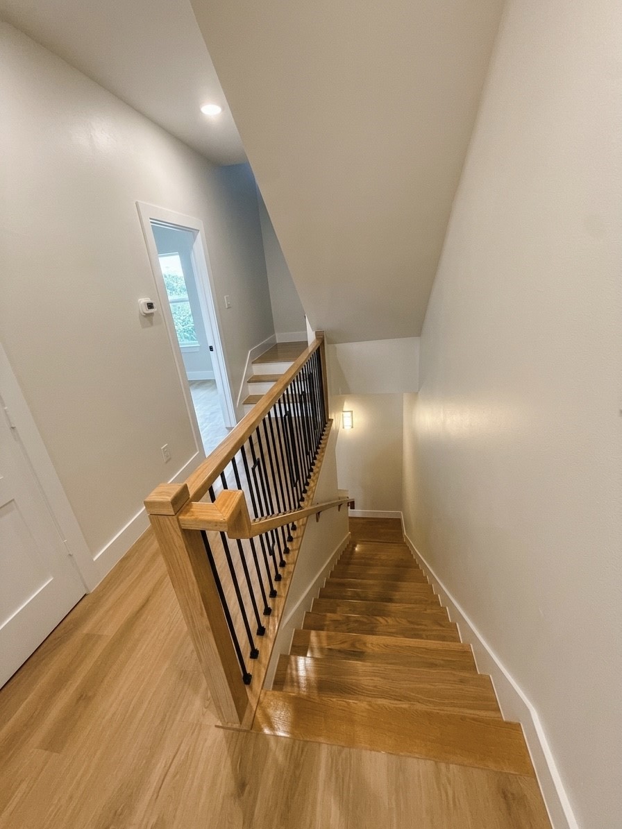 Elegant wooden staircase with sleek black railing, leading to a bright upper hallway with crisp white walls and modern lighting.