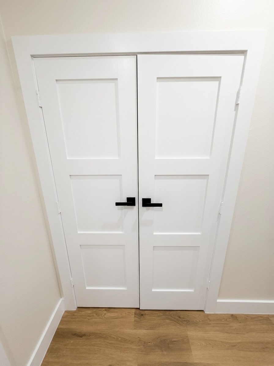 Sleek white double doors with modern black handles offer a stylish entrance to laundry room, complementing the warm engineered wood flooring.