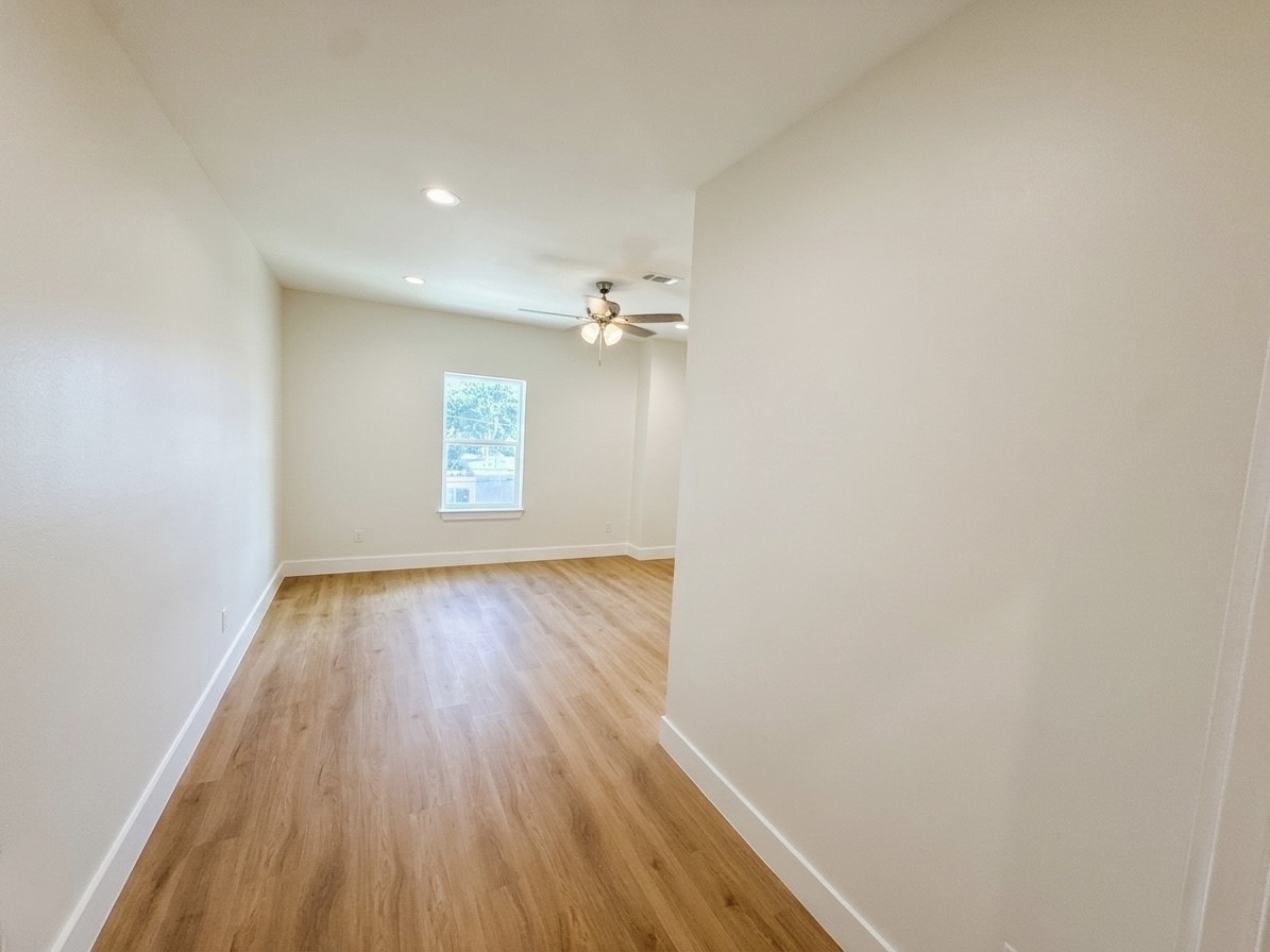 Bright and spacious room with warm engineered wood flooring, recessed lighting, and a ceiling fan, offering a cozy atmosphere. A large window provides ample natural light.