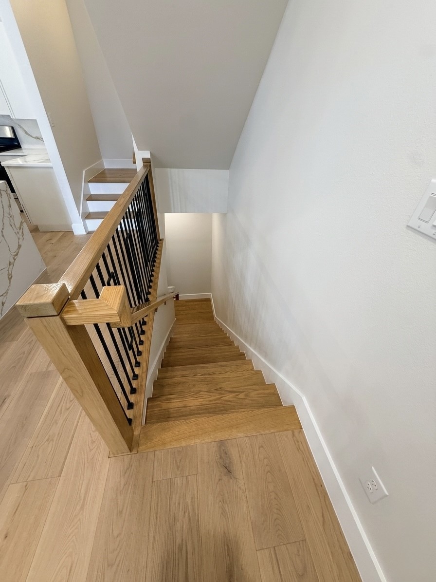 Elegant wooden staircase with sleek black railings, leading down from a bright, open space with light wood flooring.