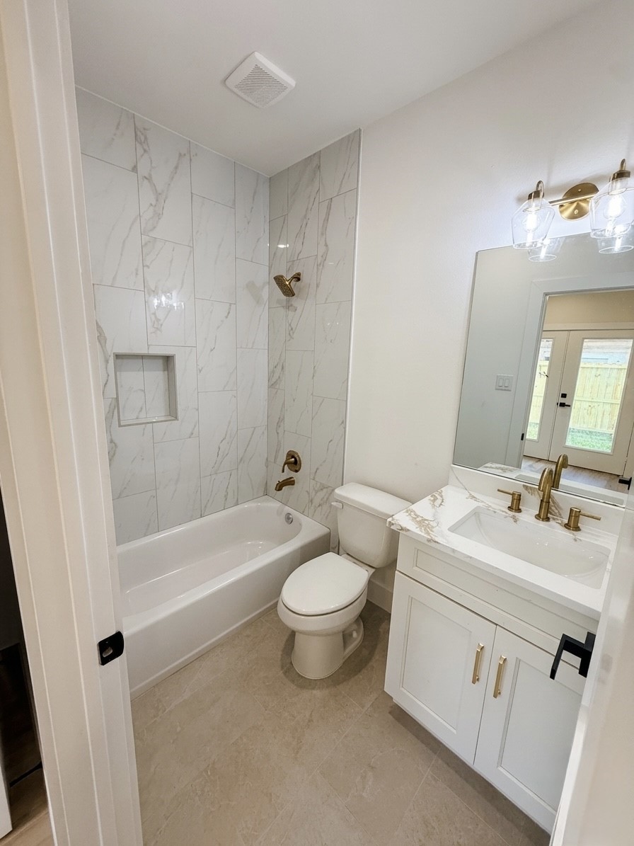 Elegant bathroom 1st floor featuring a modern tub with marble-style tile, chic brass fixtures, and a stylish vanity with ample lighting.