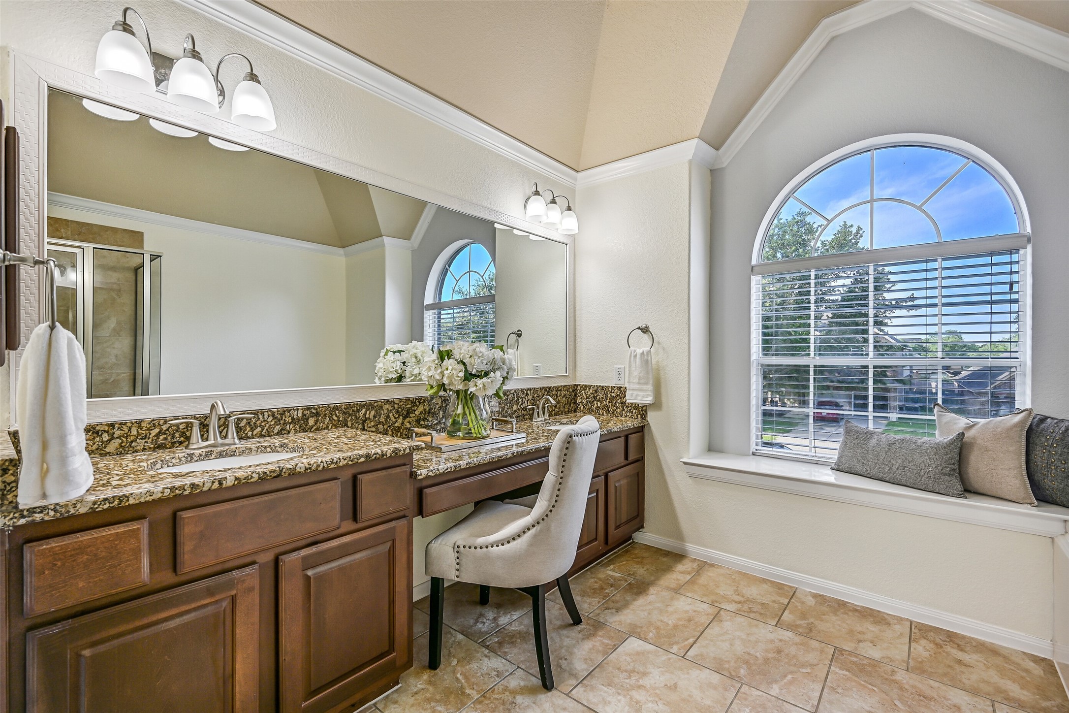 The expansive vanity offers abundant countertop space, handsome raised-panel cabinetry, dual sinks, updated lighting, and a dedicated seated dressing area. The oversized arched window and elevated ceiling details add character and create a bright, airy feel throughout the space.