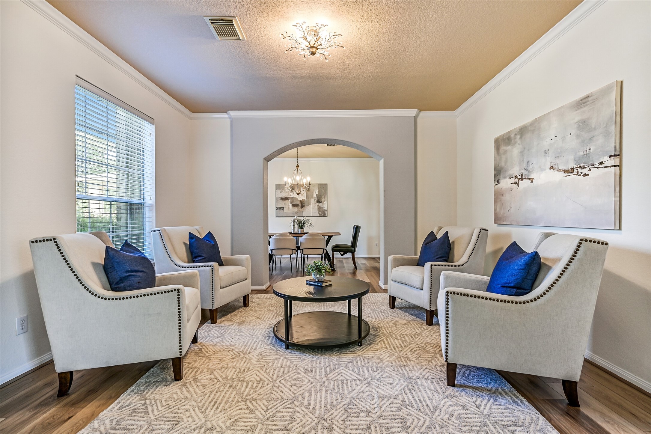 A wide arched entrance from the foyer welcomes guests into this versatile formal living room featuring fresh neutral paint, elegant crown molding, a stylish updated light fixture, and newly installed luxury vinyl plank flooring. A large front-facing window fills the room with natural light, creating an inviting space for conversation, reading, or entertaining.