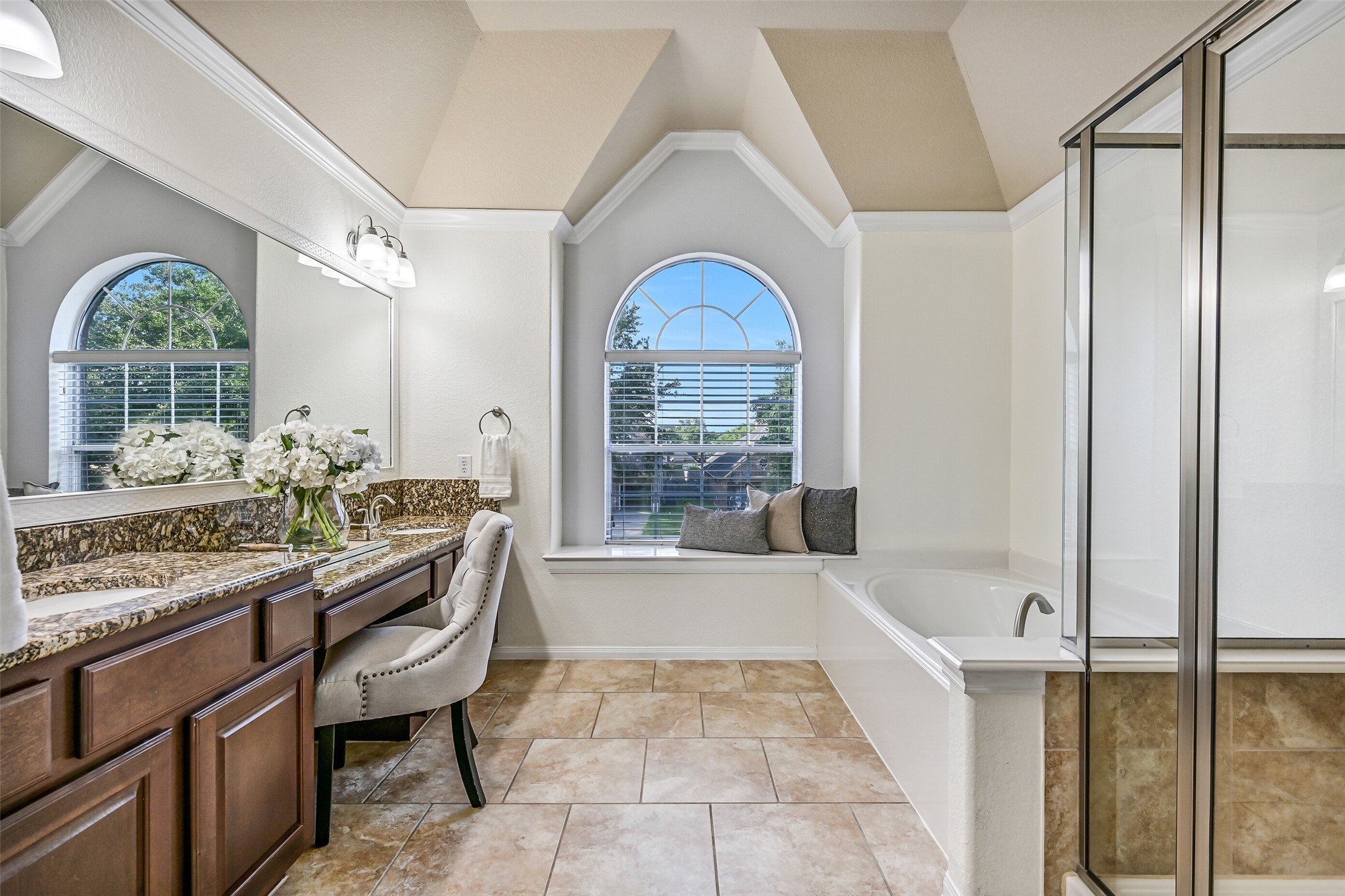 The ensuite bathroom continues the spacious feel of the primary suite with a vaulted ceiling, decorative architectural details, tile flooring, and a granite-topped dual-sink vanity. A built-in seated makeup station sits beneath a large framed mirror, while the arched window fills the room with natural light.