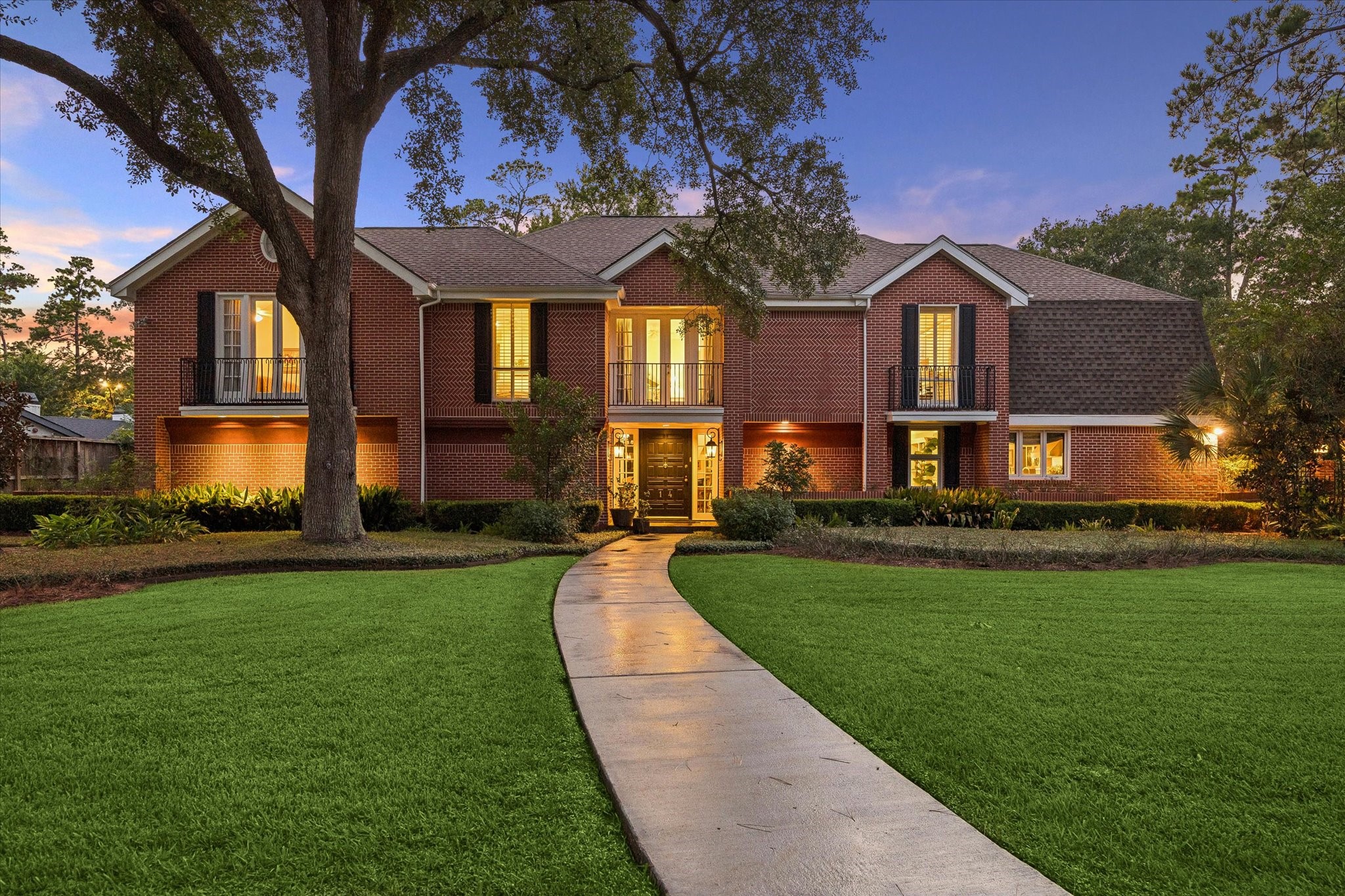 Welcome to 14 Stonegate, a stately  brick home with elegant lighting, surrounded by lush greenery and towering trees.