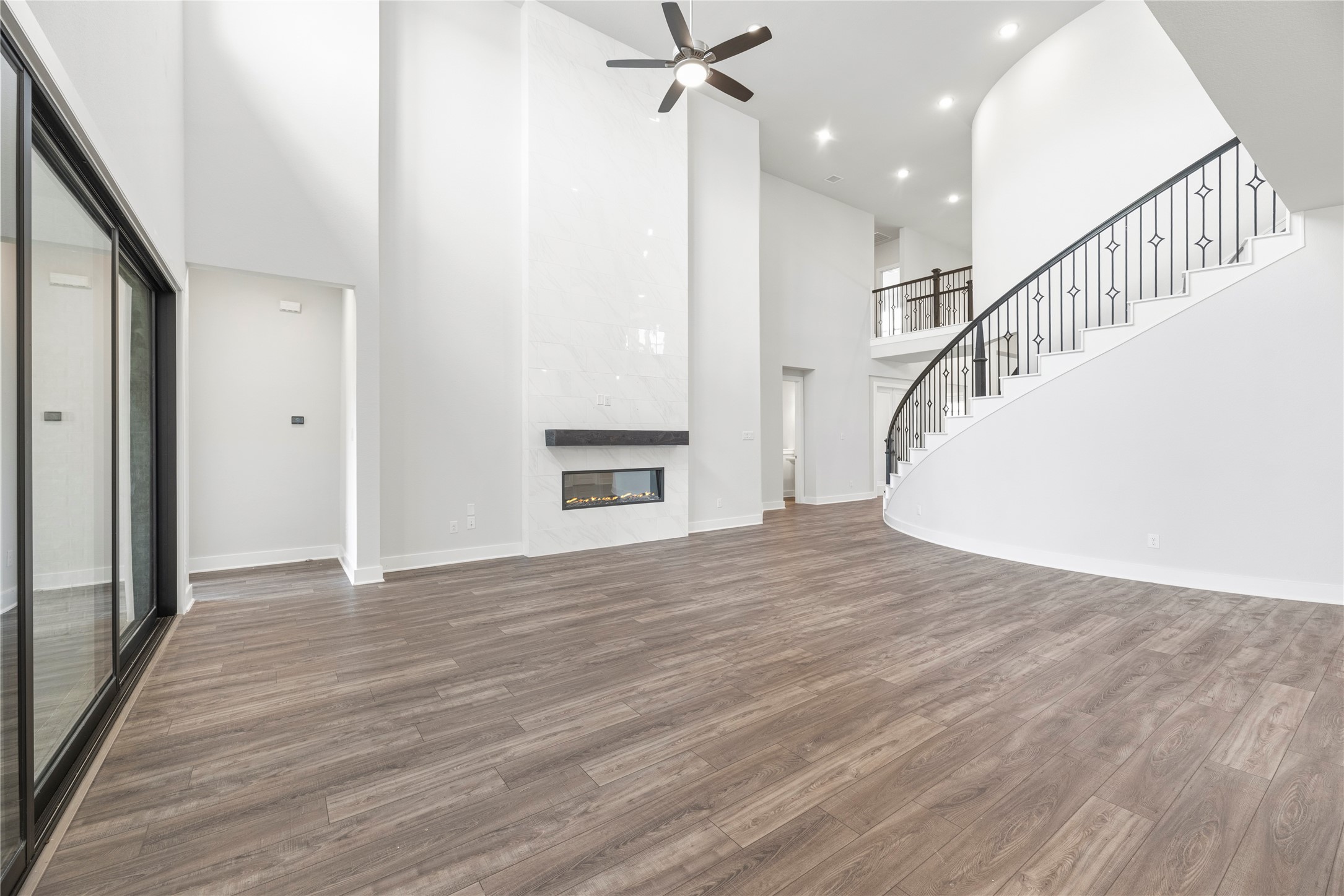Angle showcasing the sleek linear fireplace, wood-look plank flooring, grand curved staircase with wrought-iron railing, and large sliding glass patio doors.