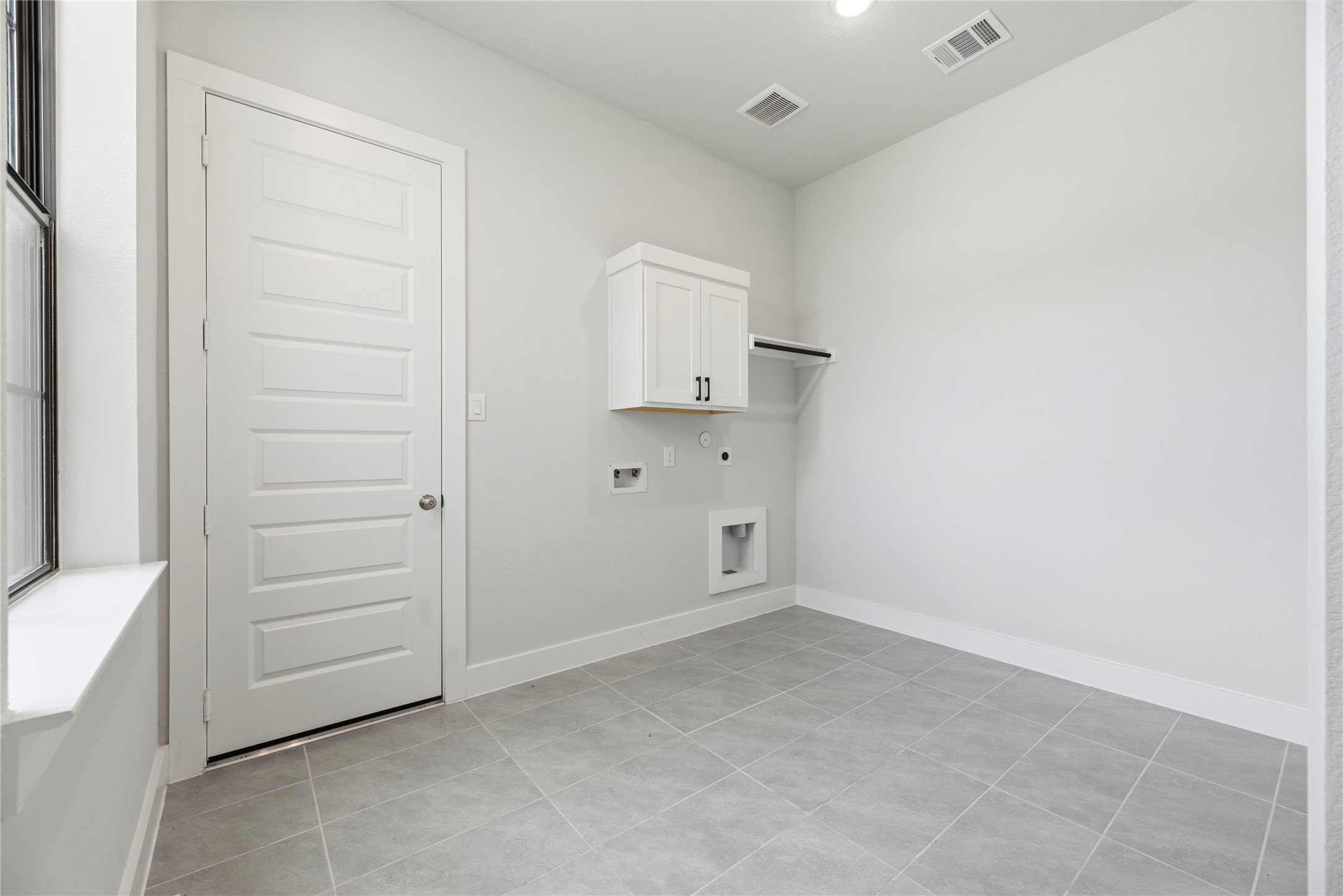 Dedicated utility room featuring easy-maintenance tile flooring, upper storage cabinetry with hanging rod, washer/dryer hookups, and natural window lighting.