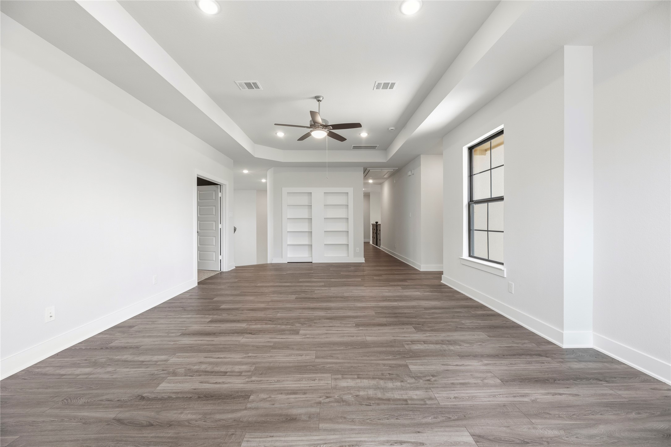 Generous game room featuring tray ceilings, wood-look flooring, and custom built-in white shelving along the rear wall.