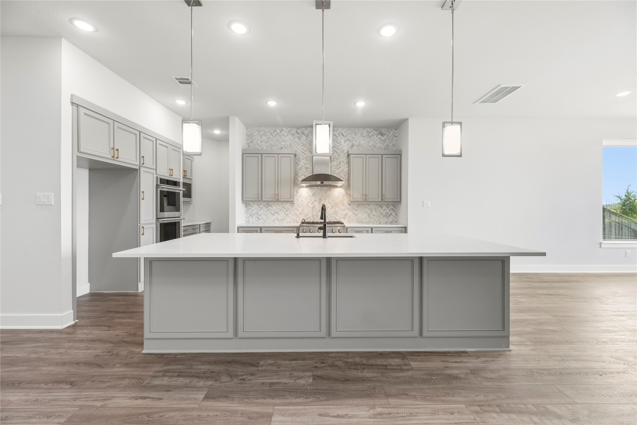 Center view of the massive prep island with built-in sink, dark hardware accents, triple glass pendant lights, and stainless steel range hood setup.