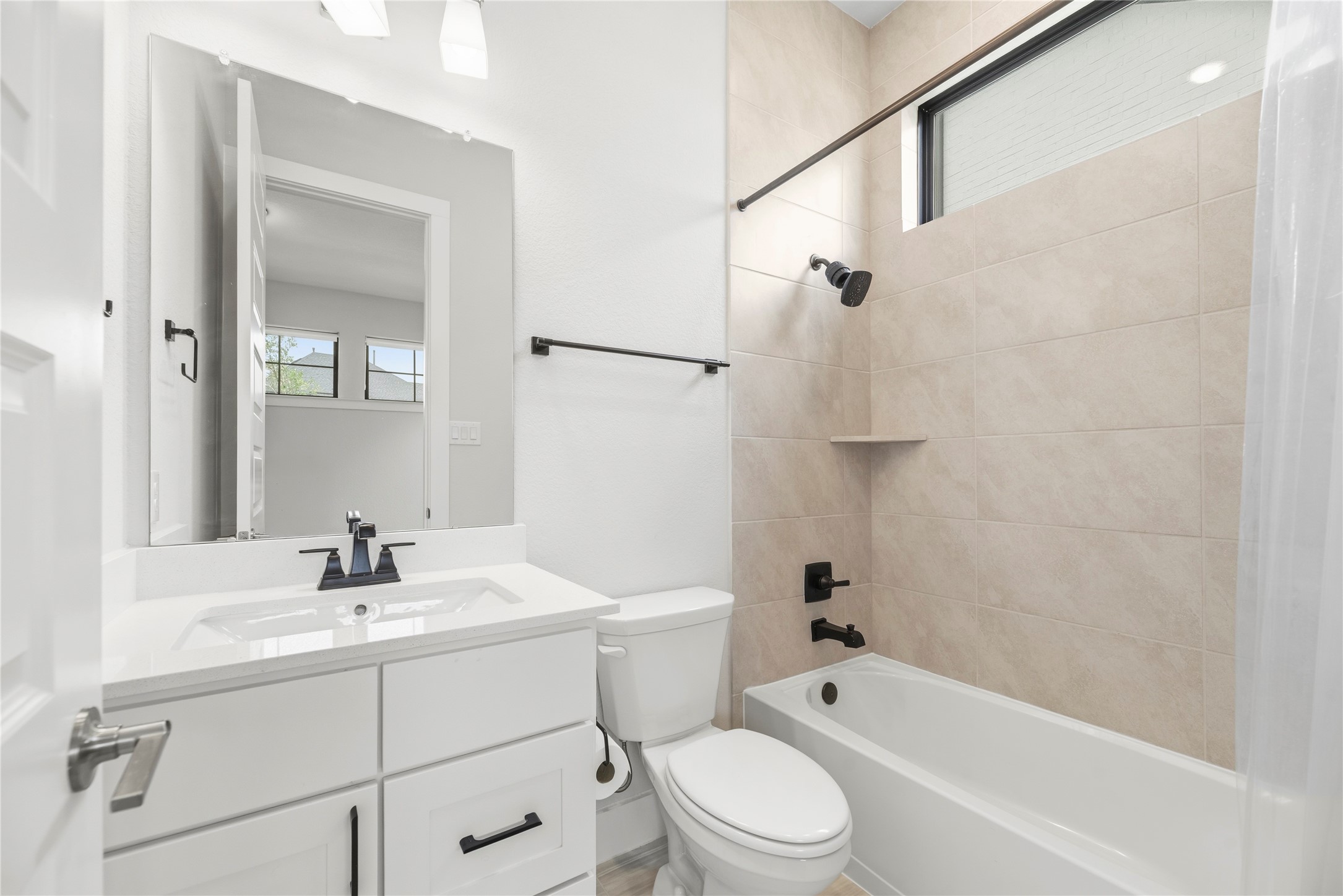Clean, modern full bath featuring a white vanity with quartz counter, dark matte fixtures, and a tiled tub/shower combo with a natural light transom window.