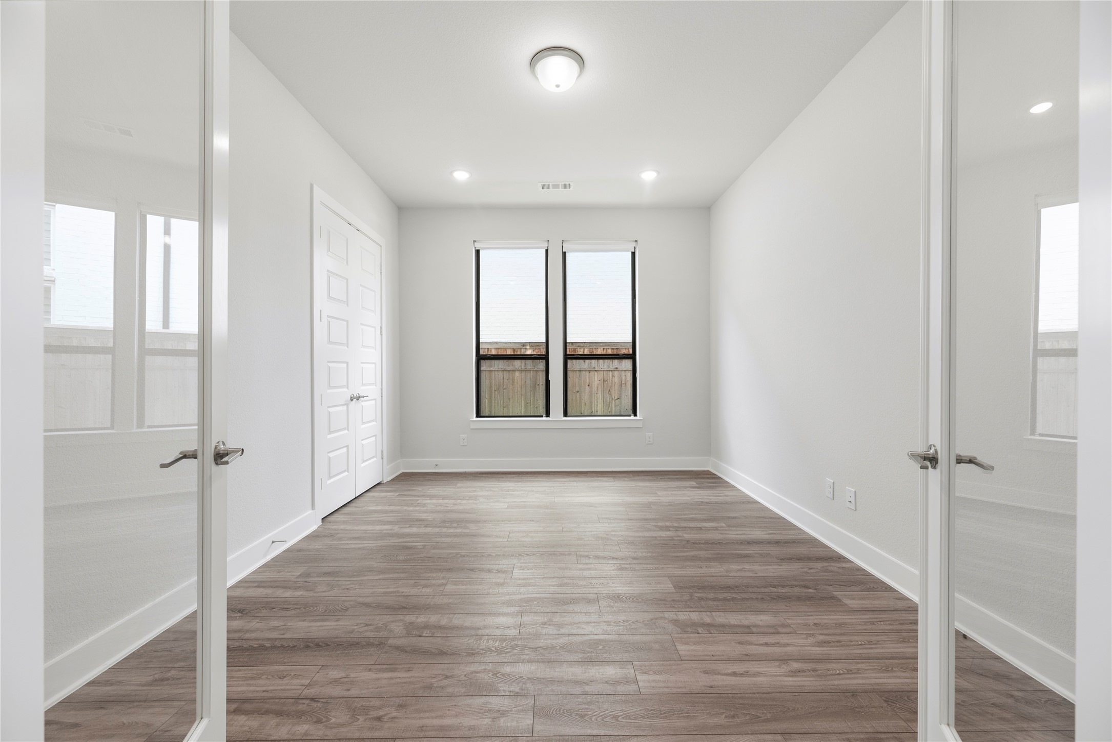 Dedicated main-floor office framed by elegant double French doors, featuring wood-look flooring, recessed lighting, and rear-facing windows.