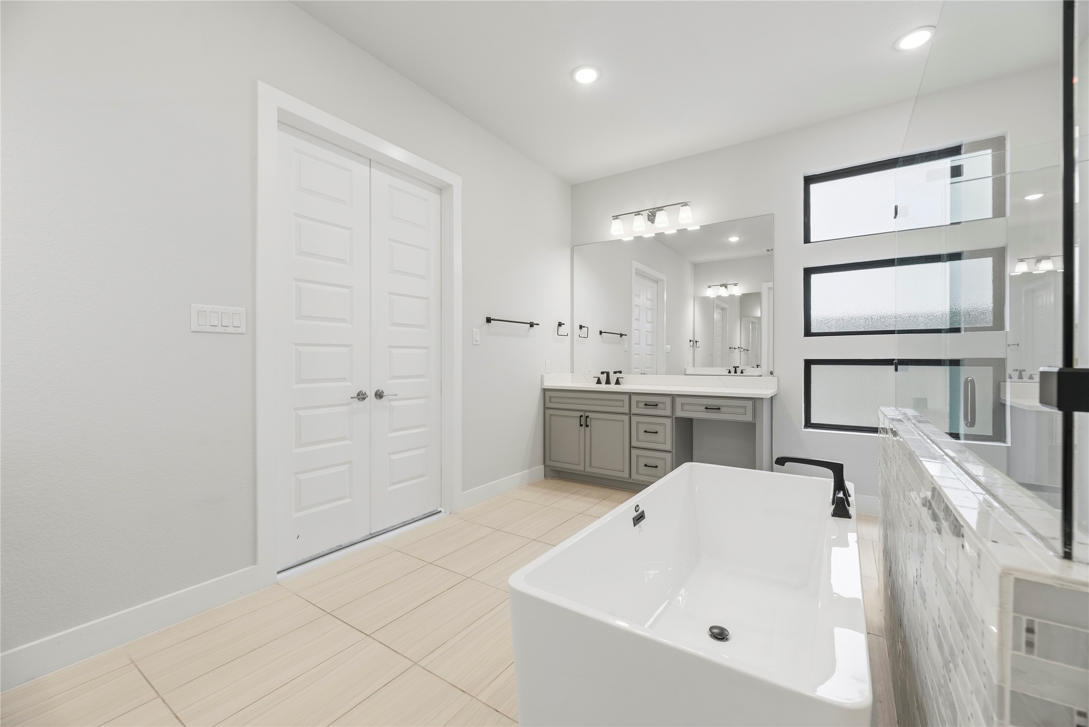 Angle highlighting the standalone tub, matte black plumbing hardware, neutral floor tile, and three stacked frosted privacy windows above the vanity.