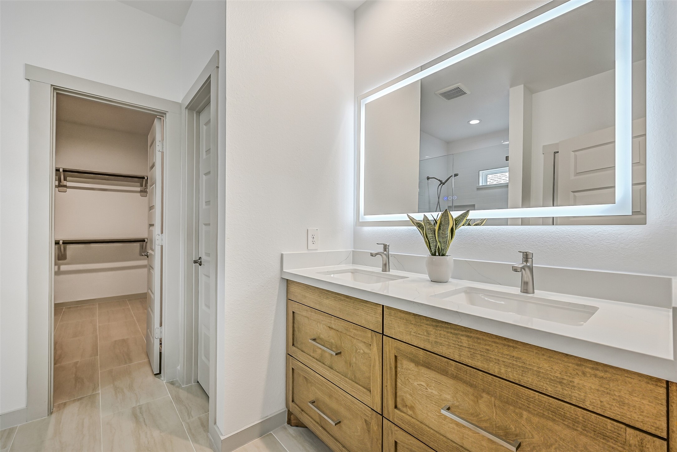 The primary bath features a long dual-sink vanity with warm wood-tone cabinetry, sleek fixtures and an impressive illuminated mirror, while an adjoining walk-in closet keeps storage conveniently close at hand.