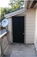 This photo shows an outdoor entrance to a home with a black door, beige siding, and a railing. There is a satellite dish attached to the railing, and trees are visible in the background.