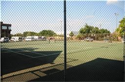 The photo shows a fenced tennis court adjacent to a parking area, surrounded by trees and a building, ideal for recreational activities.